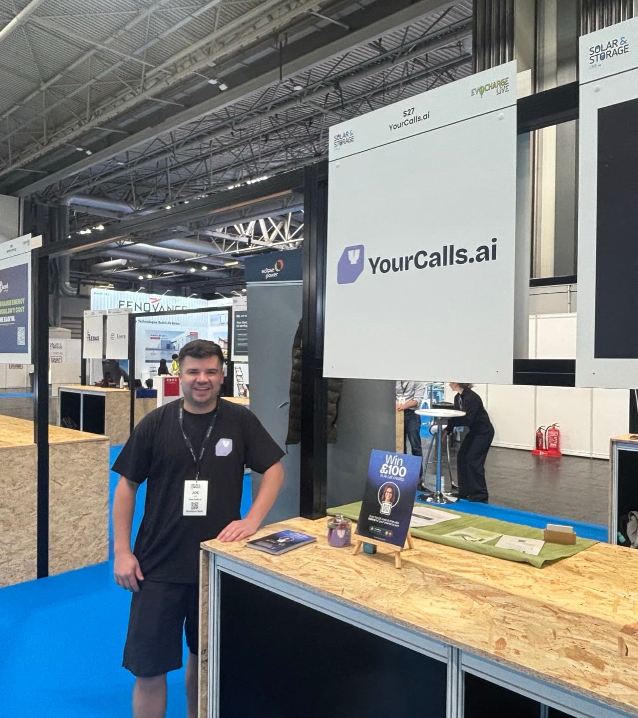The YourCalls stand at Solar & Storage Live: a white hanging sign reading S27 YourCalls.ai above the pod, with Joe standing at the counter beside it.